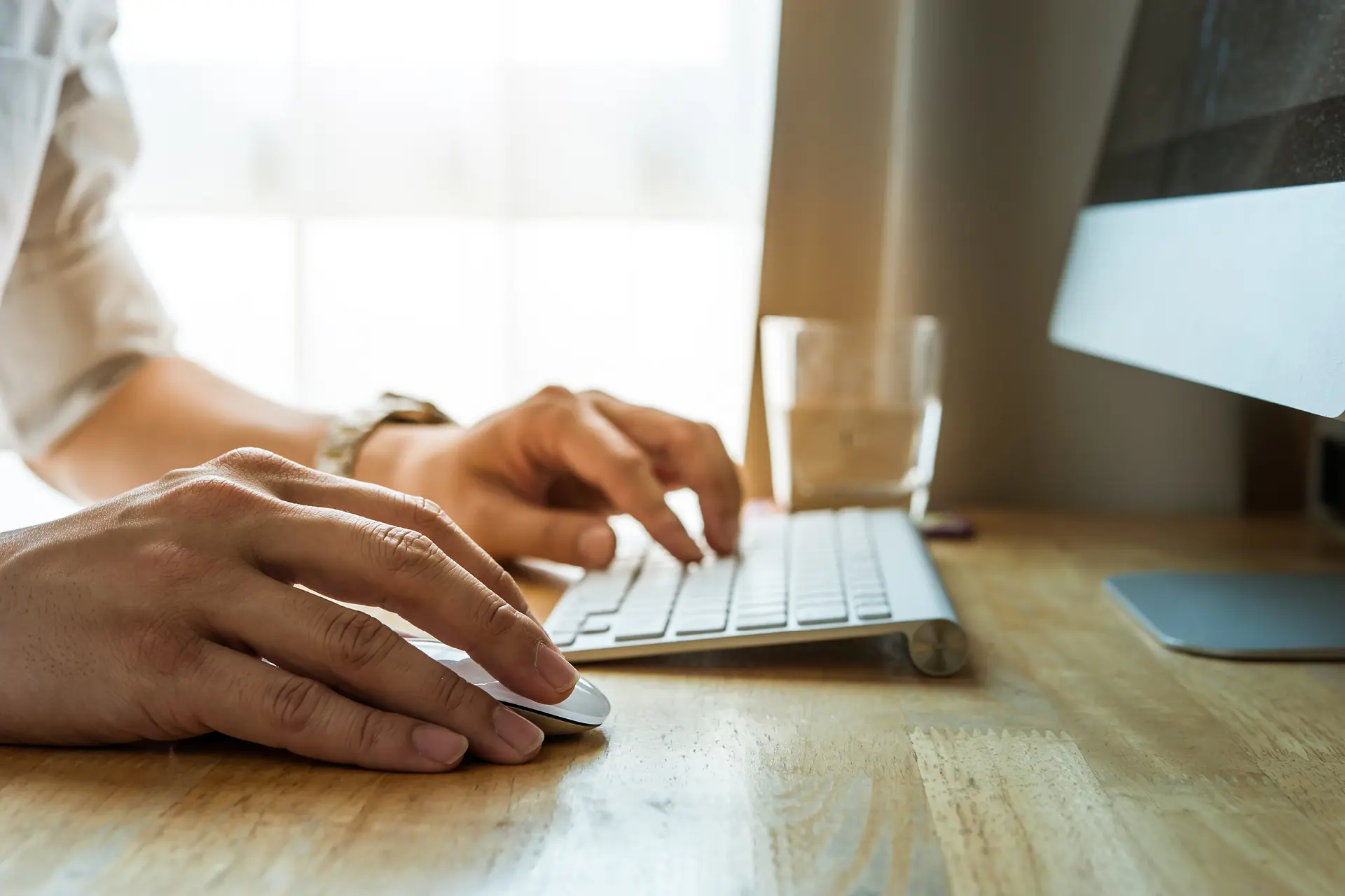 Close-up of a person using a computer mouse and keyboard at a desk near a computer monitor.